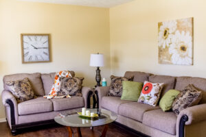 Living room staged with a beige couch along one wall and loveseat along the other wall. Wall art with a clock on one wall and flower art on another. Glass coffee table with candles in front of couches.