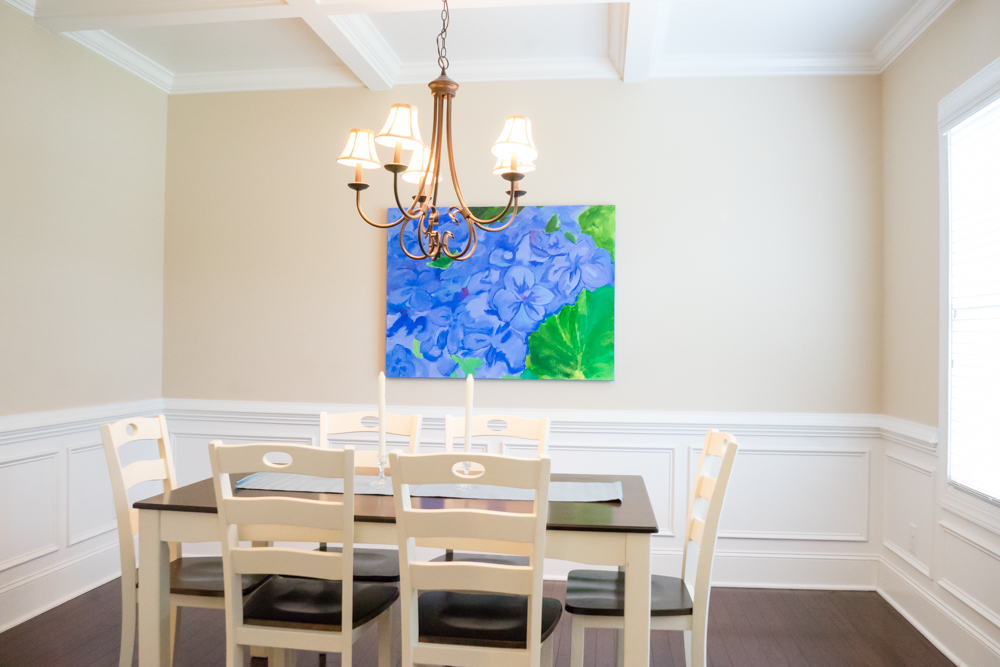 Dining room staged with farmtable in white and brown with white wooden chairs and a large canvas art piece in blue and green
